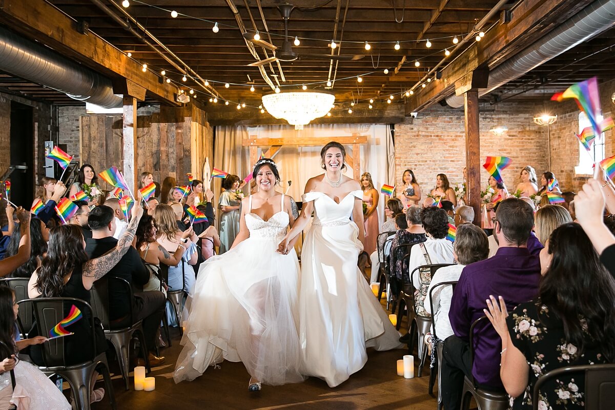 Brides exiting ceremony while guests wave pride flags