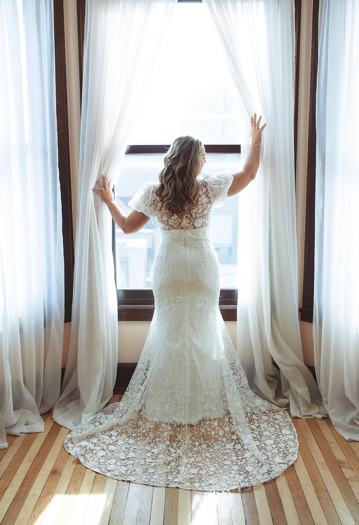 Bride pushing aside curtains while standing at window and looking out