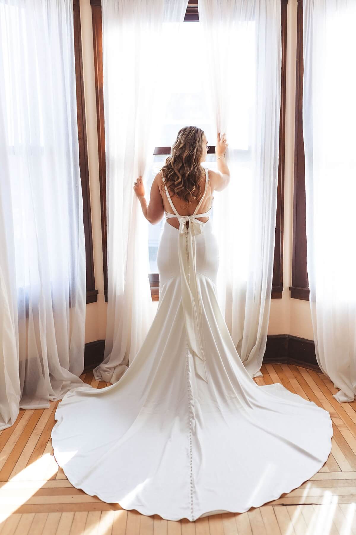 Bride wearing satin wedding dress standing in window pushing curtains aside
