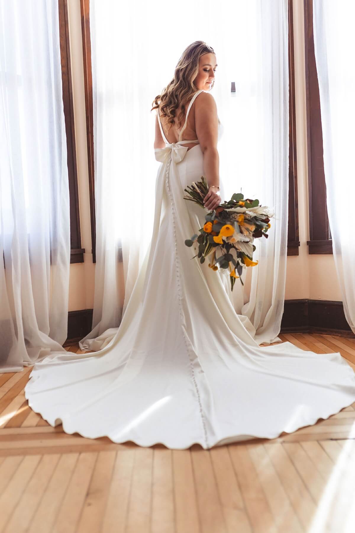 Bride wearing dress with long train holding bouquet of yellow flowers and standing in window