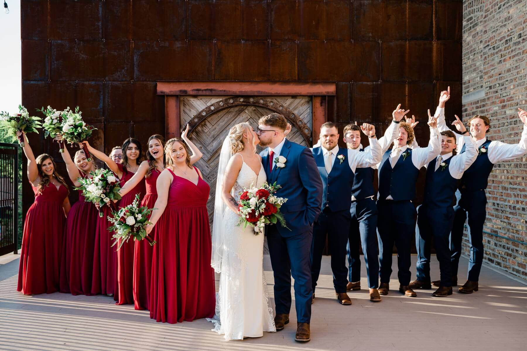 Bride and groom kissing as wedding party wearing red and blue cheer behind them