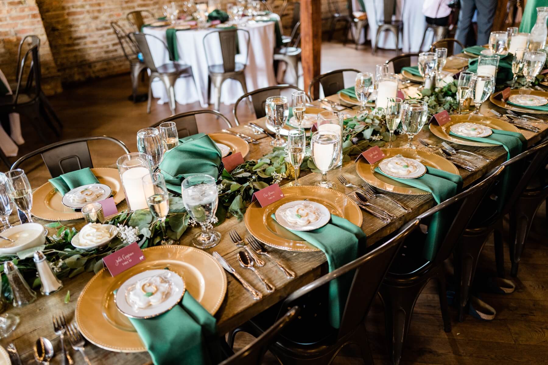 Wedding reception table with gold chargers, green napkins, and greenery